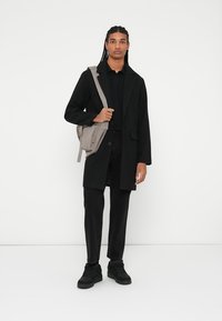 Svart lang frakk over en svart polo, kombinert med svarte bukser og sneakers. Holder en lys grå skulderveske med justerbare stropper.