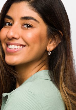Goldene Creolen zeigen ein kreisförmiges Design mit einem kleinen schwarzen Innenring. Die Person hat lange, dunkle Haare und trägt ein hellgrünes, kragenförmiges Oberteil.