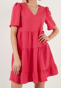 Šaty s krátkým rukávem a V výstřihem v čistě růžové barvě, s vrstveným designem a volánkovým lemem. Vyrobeno z lehkého materiálu s hladkou texturou.