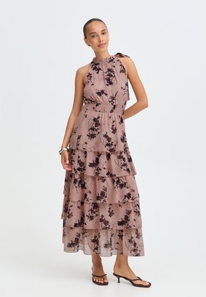 Femme portant une robe mauve sans manches à volants avec imprimé floral noir et des sandales noires à talons, debout avec une main sur la hanche.