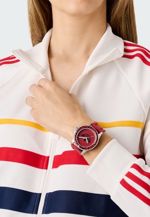 Red watch with a round face, silver accents, and a textured rubber strap. The wearer is in a white zip-up jacket with colorful stripes.