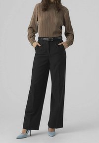 Haut long à manches plissées marron et pantalon noir taille haute, avec poches latérales, accompagné de talons aiguilles bleu clair.