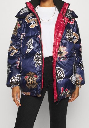 Personne portant une doudoune bleu marine avec une doublure rouge, ornée de divers écussons de style universitaire, par-dessus une chemise blanche et un jean noir.