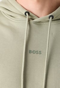 Sweat à capuche vert clair avec cordons et petit texte "BOSS" imprimé sur la poitrine, porté par une personne.