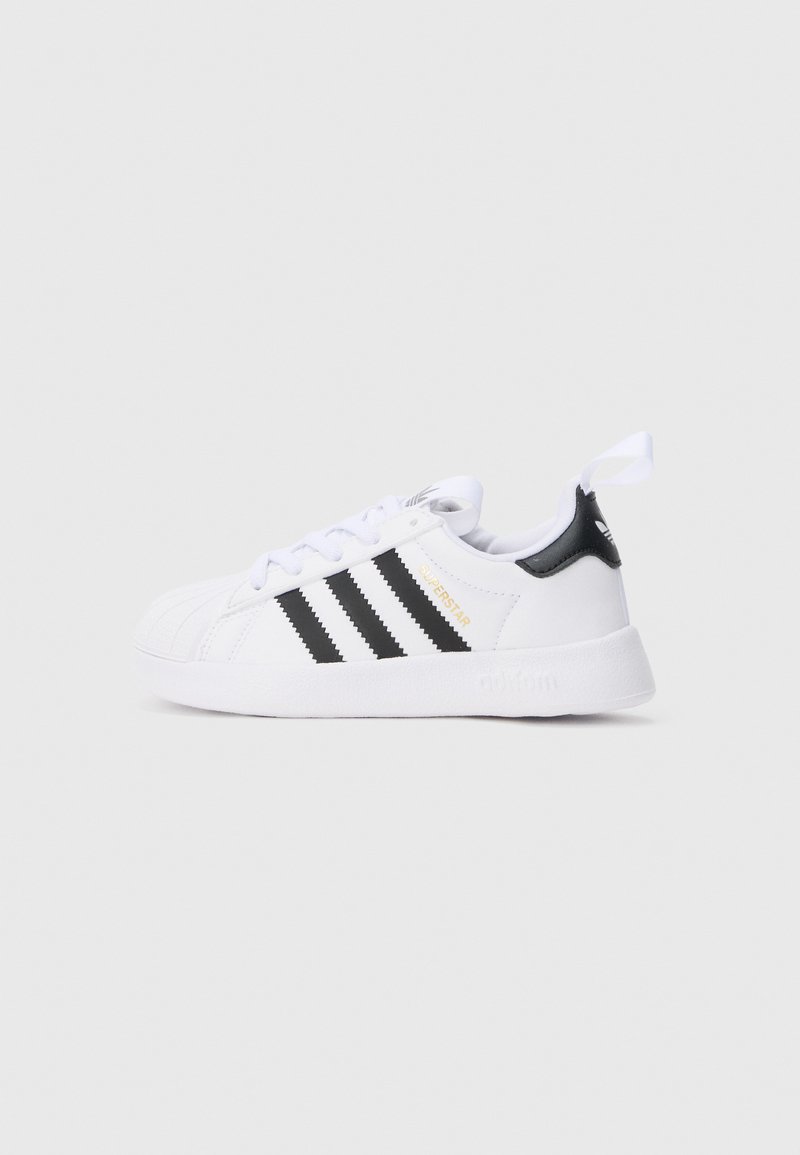 Λευκό αθλητικό παπούτσι Adidas Superstar με μαύρες ρίγες, χρυσή επιγραφή "Superstar", μαύρη γλώσσα στη φτέρνα και λευκή θηλιά τραβήγματος, πλευρική όψη σε απλό φόντο.