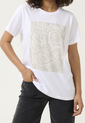 Vrouw draagt een wit T-shirt met een beige abstract bloemmotief en het geborduurde woord "FLOWERS," gecombineerd met een zwarte spijkerbroek, hand in de zak.