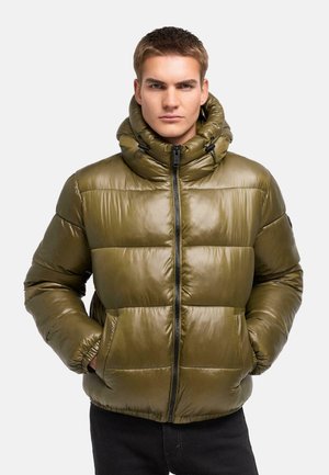 Een jonge man draagt een olijfgroene donsjas met een hoge, gevoerde kraag en heeft zijn handen in de zakken, staand tegen een effen achtergrond.