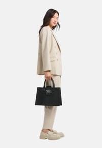 Borsa strutturata nera con forma rettangolare, dotata di manico superiore e dettaglio con logo. Il modello indossa anche un completo beige e scarpe.