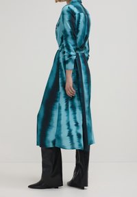 Kobieta w tealowo-czarną sukienkę midi w stylu tie-dye z długimi rękawami, w czarnych kozaczkach do kolan, stojąca bokiem na białym tle.