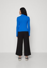 Cămașă albastră cu mânecă lungă și guler înalt, asortată cu pantaloni negri cu picioare largi și adidași gri. Materialul pare neted și ajustat.