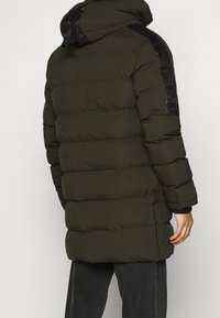 Abrigo acolchado en verde oscuro con capucha, con paneles negros en los hombros, mangas largas, corte hasta la mitad del muslo y detalles de cremalleras laterales.