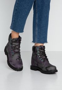 Botas de camuflaje moradas con acentos negros, que presentan un diseño de cordones, una superficie texturizada y ojales en turquesa en contraste. Suela de goma con dibujo.