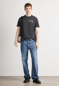 Γκρι βαμβακερό T-shirt με λογότυπο, συνδυασμένο με ανοιχτό μπλε τζιν παντελόνι. Το μοντέλο φοράει μαύρα παπούτσια και μια γκρι τσάντα χιαστί, στέκεται σε ουδέτερο φόντο.