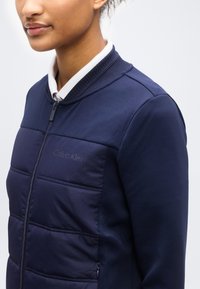 Γυναίκα που φοράει μπλε σκούρο βαμβακερό μπουφάν με φερμουάρ μπροστά και λευκό πουκάμισο με γιακά από μέσα, με το όνομα της μάρκας διακριτικά ορατό στο στήθος.