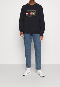 Uomo che indossa un maglione blu navy con logo oro e rosso, jeans blu e sneakers bianche alte, in piedi contro un muro bianco.