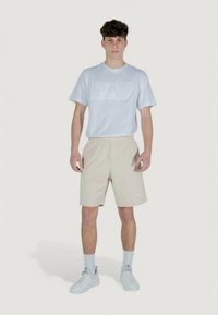 T-shirt bianco in cotone con logo "EA7" in rilievo, abbinato a pantaloni corti beige con una stampa sottile "EA7". Il modello indossa sneakers e calzini bianchi.