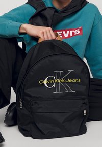 Personne portant un sweat-shirt Levi's bleu sarcelle tenant un sac à dos Calvin Klein Jeans noir par la poignée supérieure, assise en tailleur.