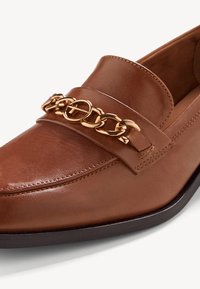 Mocassin en cuir marron avec un accent de chaîne dorée sur le dessus. Texture lisse avec des coutures détaillées et une semelle légèrement surélevée.