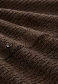 Braun strukturiertes Gewebe mit subtilen, gewellten Mustern, das einen runden Knopf mit einem genähten Loch zeigt und ein weiches, strapazierfähiges Material vermittelt.