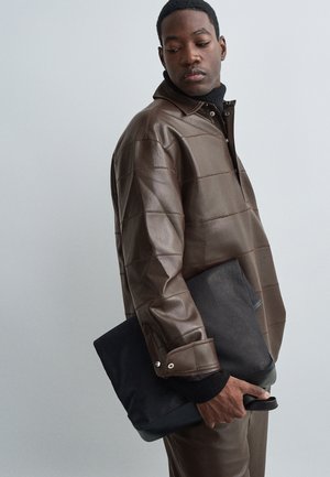 Muž v hnedej koženej bunde a nohaviciach, ktorý drží čiernu obdĺžnikovú kabelku a pozerá sa dole doľava.