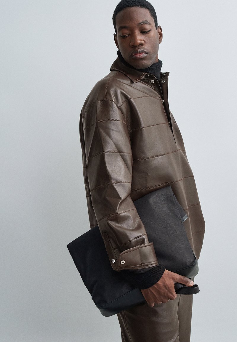 Férfi barna bőrdzsekiben és nadrágban, egy fekete téglalap alakú táskával a kezében, balra lefelé néz.