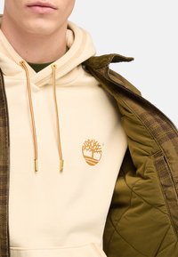 Beige hættetrøje med guldtræ-logo, sammen med en brun mønstret jakke. Hættetrøjen har snore med gyldne spidser og en glat tekstur.