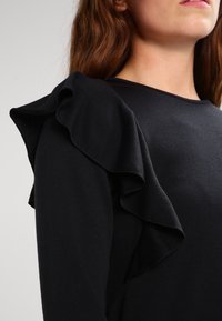 Top negro de manga larga con detalle de volantes texturizados en el hombro. Tejido suave con un acabado liso. Cuello redondo simple, corte ajustado.