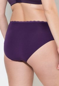 Kvinna som bär högmidjade lila underkläder och matchande bh, som visar nedre delen av ryggen och övre låren mot en enhetlig bakgrund.