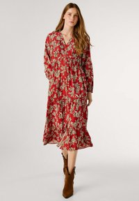 Robe maxi rouge à fleurs avec des manches longues, un devant à boutons et une taille froncée. Présente une silhouette fluide et des motifs floraux de couleur crème.
