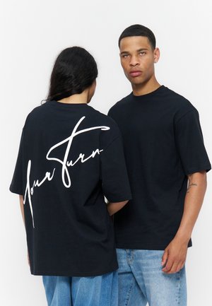 Man en vrouw in zwarte T-shirts; op de rug van de vrouw staat witte tekst op het shirt, beiden dragen losse blauwe jeans, staand tegen een effen achtergrond.