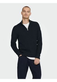 Tamno plavi pulover s polu-zip ovratnikom, rebrastom teksturom, dugim rukavima i opuštenim krojem, složen preko bijele košulje.