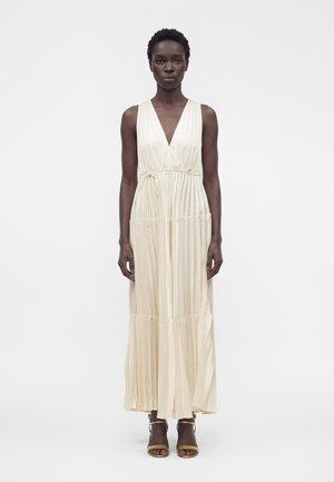 Mujer de pie, vestida con un vestido maxi plisado sin mangas de color crema, con escote en V pronunciado y cintura atada, combinado con tacones abiertos dorados.