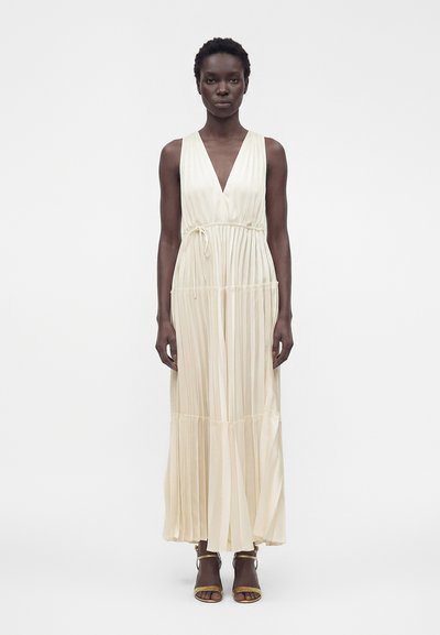 Femme debout, portant une robe longue plissée sans manches de couleur crème avec un profond décolleté en V et une taille nouée, associée à des talons ouverts dorés.
