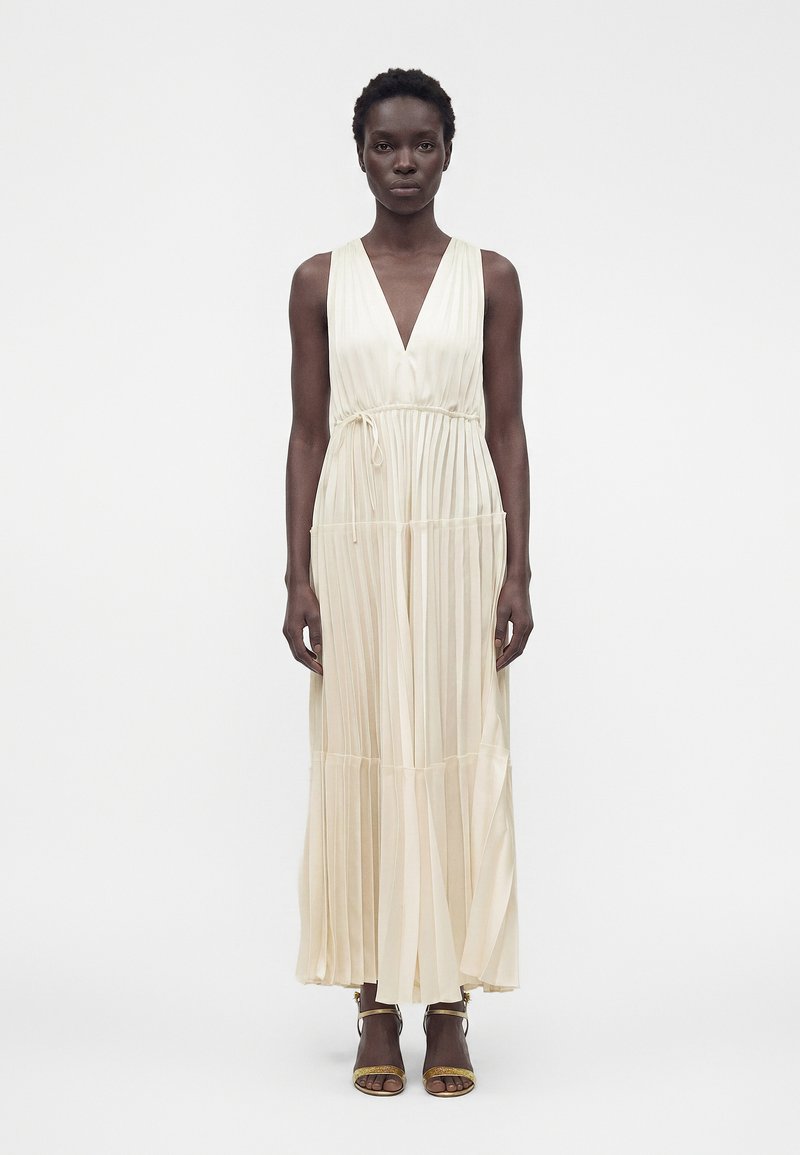 Femme debout, portant une robe longue plissée sans manches de couleur crème avec un profond décolleté en V et une taille nouée, associée à des talons ouverts dorés.