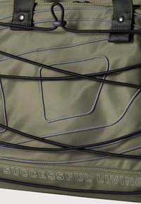 Τσάντα σε ελαιόπρασινο με μαύρα ρυθμιζόμενα κορδόνια και ραμμένο αφηρημένο σχέδιο γραμμής, με το κείμενο «ΕΠΙΤΥΧΗΜΕΝΗ ΖΩΗ» στο κάτω άκρο.