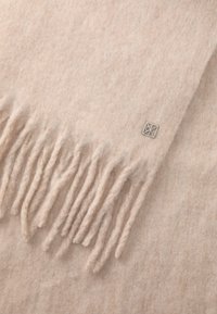 Svjetlosmeđi vuneni šal s resama i malim srebrnim logotipom, prikazujući meku, teksturiranu površinu.