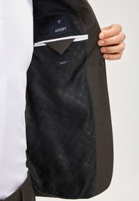 Blaser marrone scuro con una fodera nera a motivo. Presenta un'etichetta con la scritta "JOOP!" e "SLIM FIT." Dettagli di bottoni e tasche visibili.