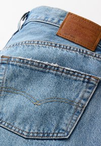 Jeans de mezclilla azul claro con un parche de cuero y costuras naranjas. Presenta un bolsillo trasero con un patrón de curva característico. Tejido texturizado.