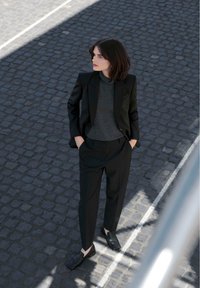 Černý přizpůsobený blazer přes šedý rolák, v kombinaci s černými kalhotami a polobotkami se čtveratou špičkou, na pozadí z kamenů.