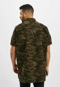 Camicia a maniche corte in camouflage verde oliva con motivi di testo ricamati, design con colletto e una vestibilità rilassata, leggermente più lunga.