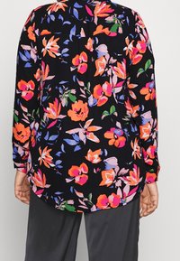 Blouse à manches longues florale noire avec des motifs vibrants de rose, orange, violet et vert ; elle présente un ourlet arrondi et des poignets boutonnés.