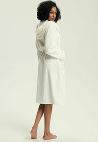 Szlafrok z kapturem z miękkiej, kremowej tkaniny o pionowej żebrowanej fakturze, z paskiem w talii i kieszeniami. Zaprojektowany z myślą o komforcie i łatwości noszenia.