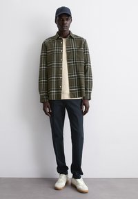 Camicia a quadri verde oliva sopra una t-shirt crema, abbinata a pantaloni scuri e sneakers bianche. Il modello indossa un berretto blu navy.