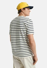 Camiseta de manga corta a rayas en blanco con rayas marinas, que presenta un escote redondo. Combinada con una gorra amarilla y marina.