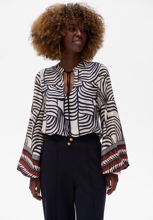 Langärmlige Bluse in Creme mit schwarzen Wellenmustern, einem Schlüsselloch-Ausschnitt und roten geometrischen Akzenten an den Ärmeln. Glattes Material.