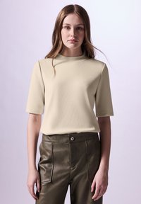 Beige, strukturovaný top s krátkým rukávem a vysokým límcem, kombinovaný s úzkými, tmavě zelenými kalhotami z umělé kůže. Jednoduchý design, minimální detaily.