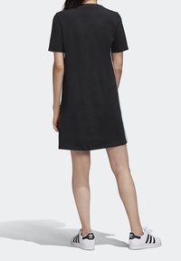 Kobieta stojąca, ubrana w czarną sukienkę z krótkim rękawem i białe trampki Adidas z czarnymi paskami, przedstawiona z tyłu na jednolitym tle.