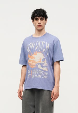 Jeune homme aux cheveux bouclés et foncés portant un t-shirt violet ample Von Dutch avec un motif de crâne en flammes et un pantalon gris, sur un fond uni.