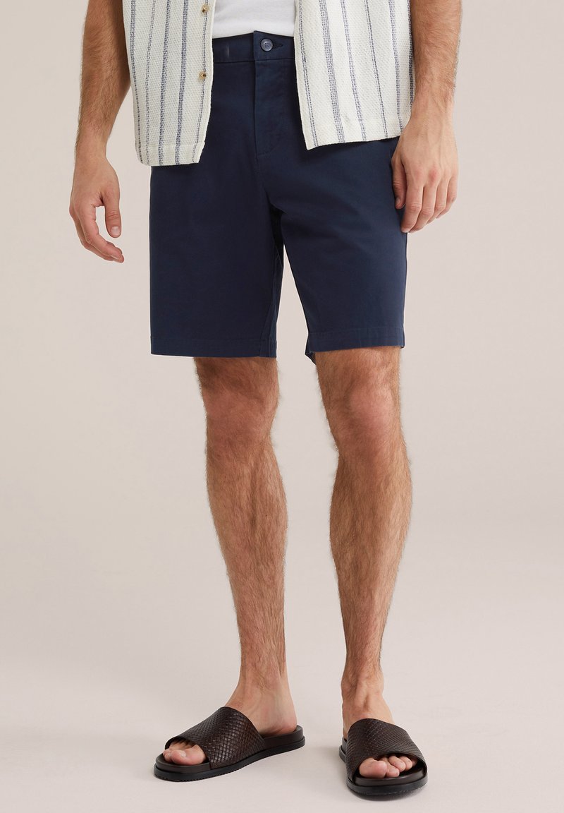 Man draagt marineblauwe korte broek, wit gestreept shirt en zwarte slippers, staand tegen een neutrale achtergrond.