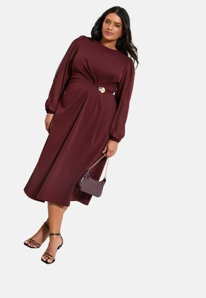 Naine, kes kannab burgundia toonis pika varrukaga midi-pikkskleiti külje pandlaga, hoides samast komplektist väikest käekotti ning mustade rihmikandadega kontsakingi.